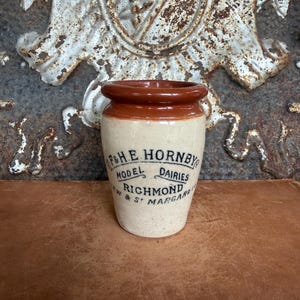 May include: A cream-colored ceramic jar with a brown glazed rim. The front features black printed text reading F & H.E. HORNBY MODEL DAIRIES RICHMOND & ST MARGARETS. The jar has a cylindrical body with a slightly flared top edge and a smooth finish.