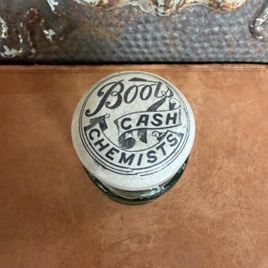 May include: A round, off-white ceramic lid with a crackled glaze finish. The top features black printed text that reads Boots Cash Chemists, arranged in a circular pattern with a central banner design. The lid is positioned on a dark glass base.