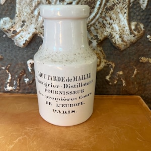 May include: A vintage white ceramic bottle with black text. The bottle reads "MOUTARDE de MAILLE Vinaigrier-Distillateur FOURNISSEUR des premières Cours DE L'EUROPE. PARIS." The bottle has a slightly speckled appearance and a rounded top.