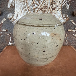 May include: A round, beige ceramic jar with a glossy, speckled glaze and horizontal dark brown bands. The vessel features a wide, short neck and a rounded body with visible surface texture, including small dark flecks and subtle imperfections.