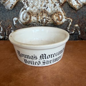 May include: A small, round, off-white ceramic bowl with a thick rim. The exterior features black text in a decorative gothic font that reads Young's Morecambe Potted Shrimps. The bowl has a smooth, glazed finish and a simple, functional design.