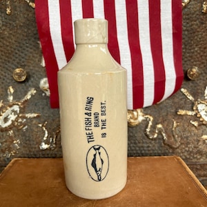 May include: A vintage, off-white ceramic bottle with a cylindrical body and a short neck. The bottle features the text "THE FISH & RING BRAND IS THE BEST" and a black and white fish illustration. An American flag is in the background.