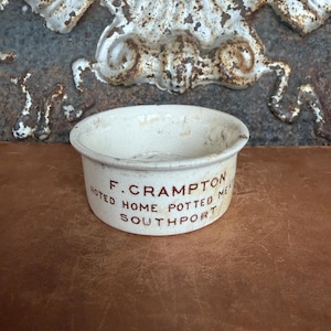 May include: A small, round, off-white ceramic pot with a visible crackled glaze finish. The exterior features brown printed text that reads F. CRAMPTON NOTED HOME POTTED MEAT SOUTHPORT. The vessel has a simple, wide-rimmed, open-top design.