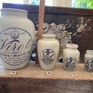 May include: Four white ceramic jars with blue and black lettering. The jars are labelled "Virol, A Preparation of Bone-Marrow, An Ideal Fat Food for Children & Invalids." The jars are arranged in a row from largest to smallest on a wooden surface.