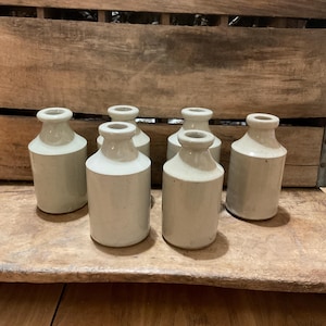 May include: Six small, white ceramic bottles with a textured surface. The bottles are arranged in two rows of three on a wooden surface.