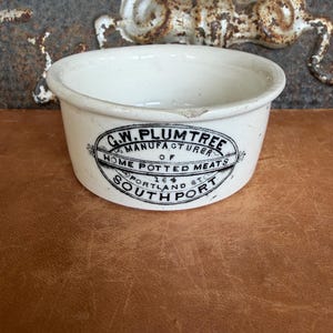 May include: A white ceramic pot with a black printed oval label that reads G.W. PLUMTREE MANUFACTURER OF HOME POTTED MEATS 164 PORTLAND ST SOUTH PORT. The round container features a slightly flared rim and a small chip on the top edge.