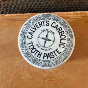 May include: A white ceramic lid with black text and floral detailing. The text reads "CALVERT'S CARBOLIC TOOTH PASTE" with "TRADE MARK" and "ONE SHILLING" also visible. The lid is circular and sits on a brown surface.