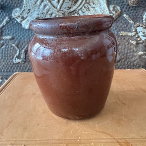 May include: A small, antique brown ceramic pot with a rounded body and a slightly flared rim. The pot has a glossy finish and appears to be handmade. It sits on a vintage book, suggesting a rustic or vintage aesthetic.