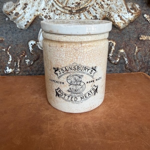 May include: A cylindrical ceramic jar with a cream-coloured crackle glaze finish and a matching lid. The front features black printed text reading J. SAINSBURY'S SUPERIOR HOME MADE POTTED MEATS with a central S logo and decorative banners.