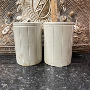 May include: Two off-white, cylindrical stoneware crocks with vertical ribbed detailing. The crocks are displayed on a dark, speckled surface, with a decorative metal backdrop. The crocks are approximately 6 inches tall.