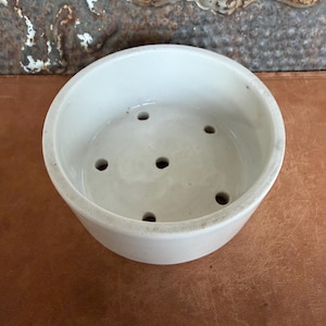 May include: A round, off-white ceramic bowl featuring a smooth interior surface with five evenly spaced drainage holes at the bottom. The bowl has a simple, cylindrical shape with a thick rim and a clean, unadorned exterior finish.