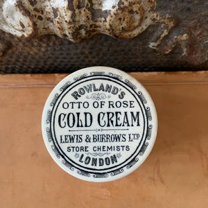 May include: A round, white container of Rowland's Otto of Rose Cold Cream. The container has black text that reads "LEWIS & BURROWS LTD STORE CHEMISTS LONDON". The container has a decorative border.