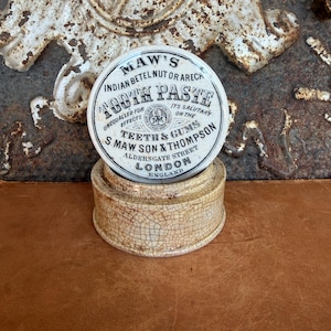 May include: A round, cream-colored ceramic pot with a lid featuring black printed text that reads Maw's Indian Betel Nut or Areca Tooth Paste, S. Maw Son & Thompson, Aldersgate Street, London, England, with a visible crackle glaze finish.