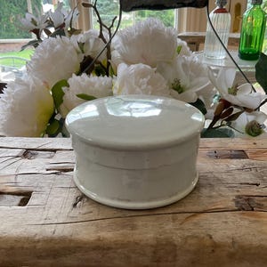 May include: A white, round, ceramic container with a lid. The container is sitting on a rustic wooden surface, with a handle in the background. White flowers and glass bottles are also visible.
