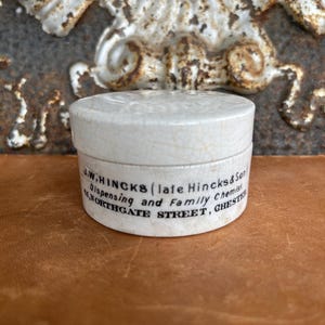 May include: A small, round, white ceramic container with a lid. The container has a cracked, aged appearance. Text on the side reads "J.W. HINCKS (late Hincks & Son) Dispensing and Family Chemist, 61, NORTHGATE STREET, CHESTER."