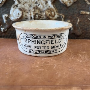 Peut inclure: Un petit bol en céramique blanc cassé avec un bord noir et du texte. Le texte indique "Horrocks & Watson SPRINGFIELD HOME POTTED MEATS SOUTHPORT" dans une police vintage. Le bol est rond avec un bord légèrement évasé.
