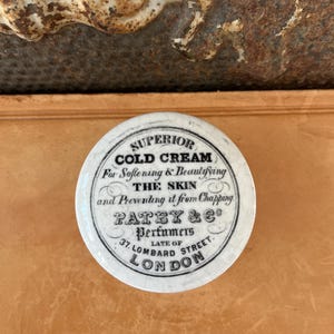 May include: A round, white ceramic jar lid with black text. The text reads "SUPERIOR COLD CREAM" and describes the product as softening and beautifying the skin. The lid also features the name "PATEY & Co, Perfumers, 37 Lombard Street, London."