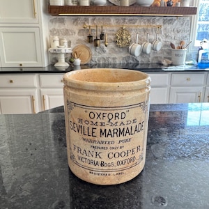 May include: A vintage, beige ceramic jar with the text "OXFORD HOME-MADE SEVILLE MARMALADE" in black lettering. The jar has a wide opening and is sitting on a dark countertop. The background shows a kitchen setting.