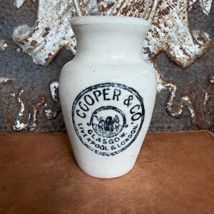 May include: A cream-colored ceramic stoneware jar with a flared rim and a rounded body. The front features a black circular stamp with the text COOPER & CO. GLASGOW & LONDON and a central emblem. The surface has a speckled, matte texture.