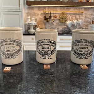 May include: Three antique James Keiller & Sons Dundee Marmalade stoneware jars. The jars are off-white with black text and a decorative border. Each jar has a different lettered tile in front of it.