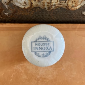 May include: A round, white ceramic soap dish with a blue floral design and the words "MOUSSE INNOXA" in a rectangular frame. The dish has a ribbed texture and is set against a warm, neutral background.