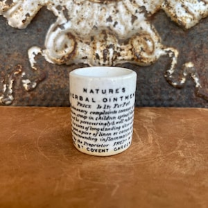 May include: A small, cylindrical, white ceramic container with black text. The text reads "NATURES HERBAL OINTMENT" and lists various ailments. The container is on a brown surface with a decorative metal background.