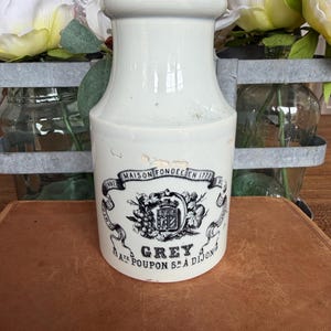 May include: A white ceramic crock with a cylindrical body and a flared rim. The front features a black printed graphic with the text Maison Fondée En 1777, Grey Poupon S.A. Dijon, and a central crest with grapes and a shield design.