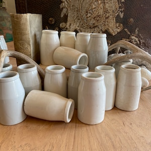 May include: A collection of antique, off-white ceramic jars of varying heights and shapes. Some are upright, others are on their sides. The jars are arranged on a wooden surface, with a weathered book and a deer antler in the background.