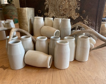 May include: A collection of antique, off-white ceramic jars of varying heights and shapes. Some are upright, others are on their sides. The jars are arranged on a wooden surface, with a weathered book and a deer antler in the background.