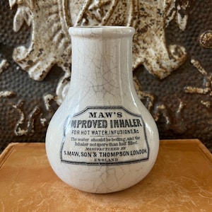 May include: A vintage white ceramic inhaler with a bulbous body and a narrow neck. The inhaler features a label with black text that reads "MAW'S IMPROVED INHALER" and instructions. The ceramic has a cracked, aged appearance.