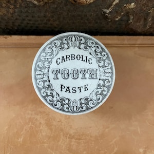 May include: A round, antique container lid with the words "CARBOLIC TOOTH PASTE" in black text. The lid has a decorative, swirling border in black and white. The background is a warm, neutral color.