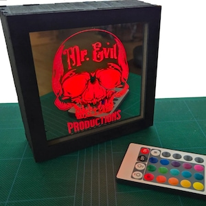 Może przedstawiać: Czarne drewniane pudełko ze szklanym frontem, na którym widnieje czerwona czaszka z napisem "Mr. Evil Productions". Na pierwszym planie znajduje się pilot z kolorowymi diodami LED.