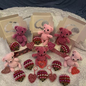 May include: A selection of handmade crochet items, including pink teddy bears, heart-shaped ornaments, and hedgehog-shaped decorations. Some bears are in decorative boxes. The items are in shades of pink and red, ideal for Valentine's Day or a similar occasion.