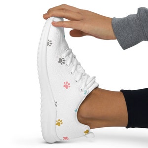 May include: White athletic shoe with colorful paw print pattern. The shoe has white laces and a white sole. The paw prints are in shades of pink, blue, yellow, and gray. The shoe is being held by a person wearing a gray sweatshirt and black leggings.