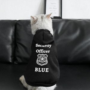 Könnte beinhalten: Eine weiße Katze trägt einen schwarzen Kapuzenpullover mit dem Text "Security Officer BLUE" und einer Abzeichengrafik.