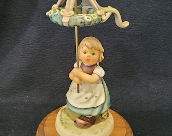 Może przedstawiać: Porcelanowa figurka dziewczynki trzymającej parasol w kształcie wieńca. Figurka stoi na drewnianej podstawie z złotą tabliczką z napisem "Sister Maria Innocentia Hummel Commemorative Edition 2001".