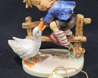 May include: A porcelain figurine of a boy in a green hat and blue jacket reaching over a fence to a white goose. The figurine has a gold tag with the number "50" on it.