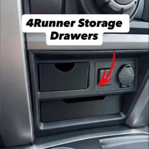 May include: Black plastic 4Runner Storage Drawers installed in a vehicle centre console. The unit features two rectangular storage compartments with cutouts for access, positioned next to a square button and a circular power outlet port.