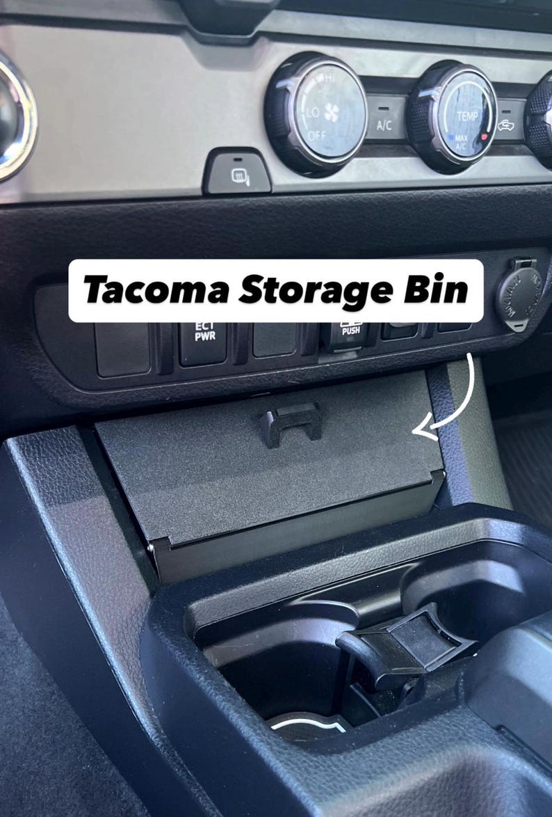 May include: Black Tacoma Storage Bin installed in a vehicle's dashboard. The bin is rectangular and has a hinged lid. The dashboard also features climate control knobs, power outlets, and other controls.