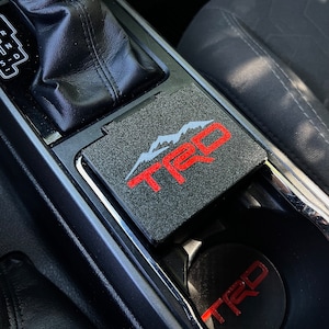 Puede incluir: Interior de coche negro con un logotipo TRD sobre una superficie texturizada. El logotipo TRD rojo presenta un diseño de cordillera. Un posavasos TRD a juego está en el portavasos. La palanca de cambios es visible en el fondo.