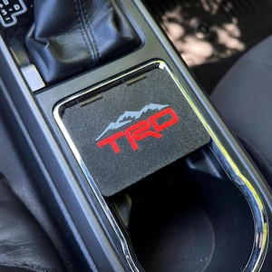 May include: A black car console cup holder insert with a red and white TRD logo and mountain graphic. The insert is placed in a car's center console, next to the gear shift.