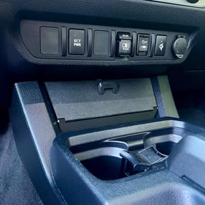 May include: A close-up of a car's dashboard, featuring a black console with various buttons and a USB port. The buttons are labeled with text such as "ECT PWR" and "REAR PUSH". A wireless charging symbol is also visible.
