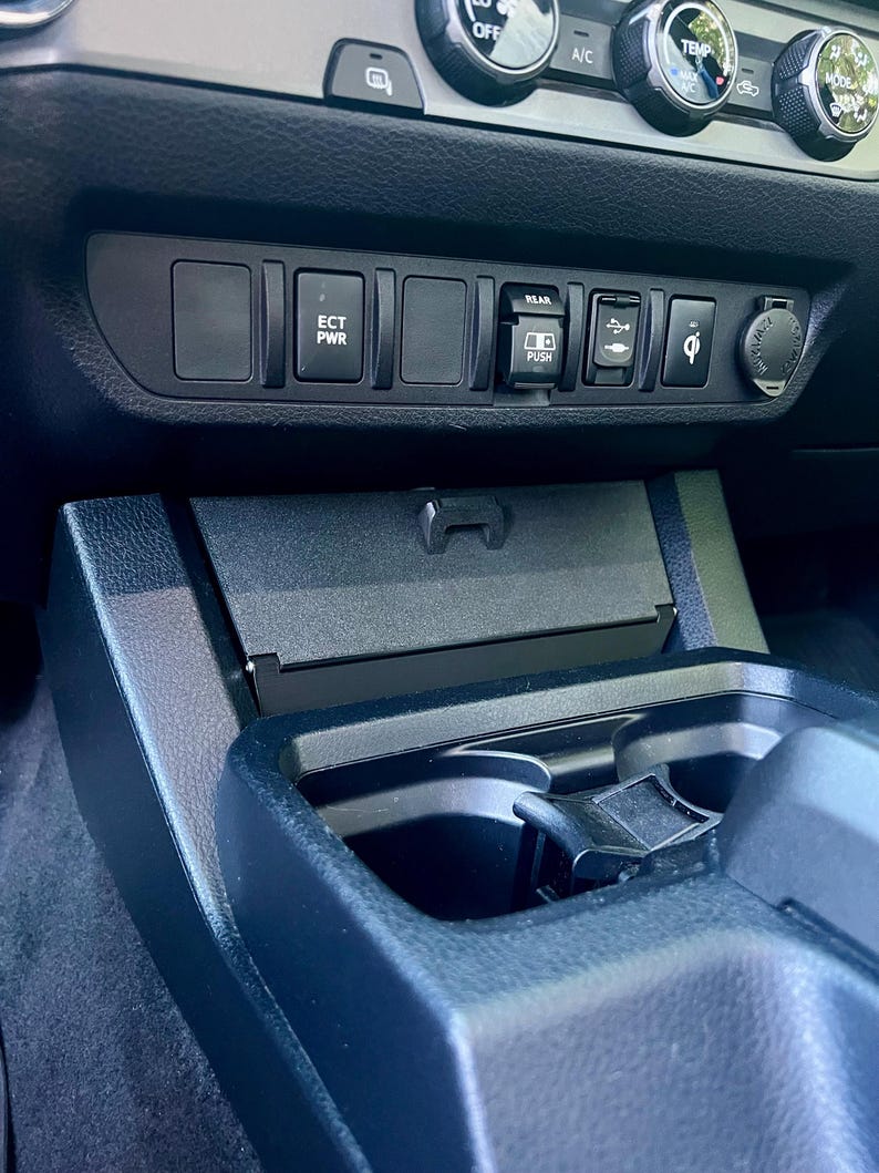 May include: A close-up of a car's dashboard, featuring a black console with various buttons and a USB port. The buttons are labeled with text such as "ECT PWR" and "REAR PUSH". A wireless charging symbol is also visible.