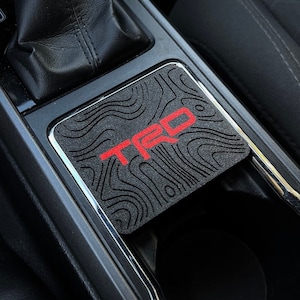 May include: A black car cup holder with a black and gray textured mat with the word "TRD" in red lettering.