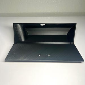 May include: A black, open-top storage box with a hinged lid. The box is made of a textured material and has a rectangular shape. Two small silver-colored rivets are visible on the lid. The box is set against a white background.