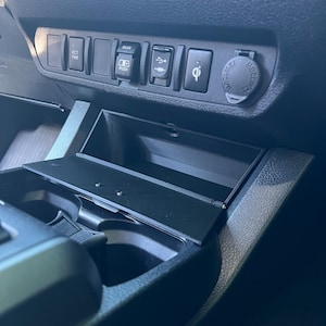 May include: A close-up of a car's interior console, featuring a black storage compartment with an open lid. The console includes buttons labeled "ECT PWR" and "REAR PUSH", along with a USB port, a wireless charging symbol, and a 12V power outlet.