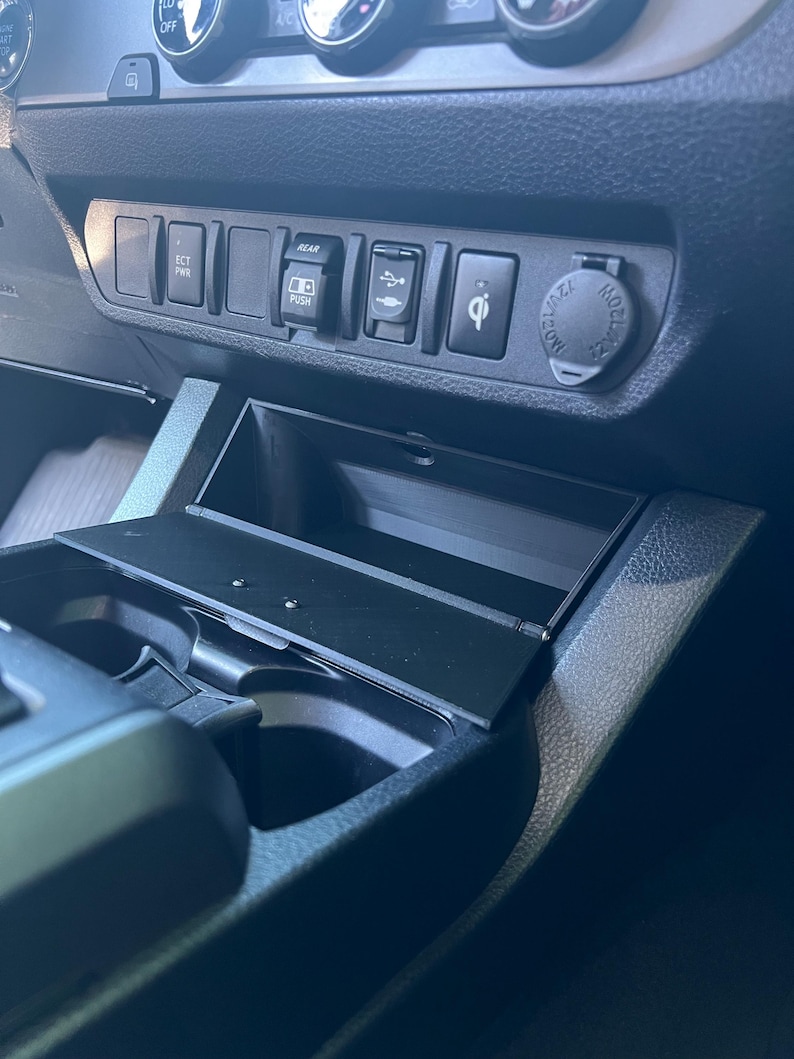 May include: A close-up of a car's interior console, featuring a black storage compartment with an open lid. The console includes buttons labeled "ECT PWR" and "REAR PUSH", along with a USB port, a wireless charging symbol, and a 12V power outlet.