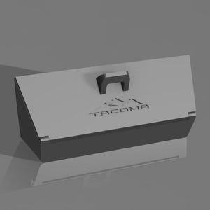 May include: A gray rectangular box with a black handle and the word "TACOMA" printed on the lid. The box has a mountain graphic above the text. The box is on a gray surface.