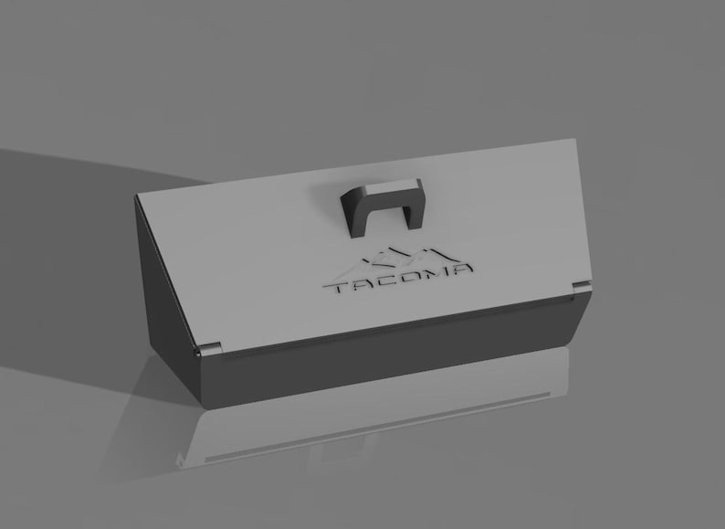 May include: A gray rectangular box with a black handle and the word "TACOMA" printed on the lid. The box has a mountain graphic above the text. The box is on a gray surface.