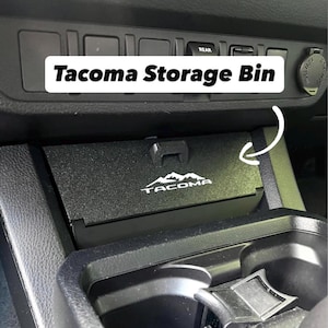 May include: Black Tacoma Storage Bin with the Tacoma logo. The bin is installed in a vehicle's dashboard, near the center console. The text "Tacoma Storage Bin" is displayed in a white rectangle above the bin.